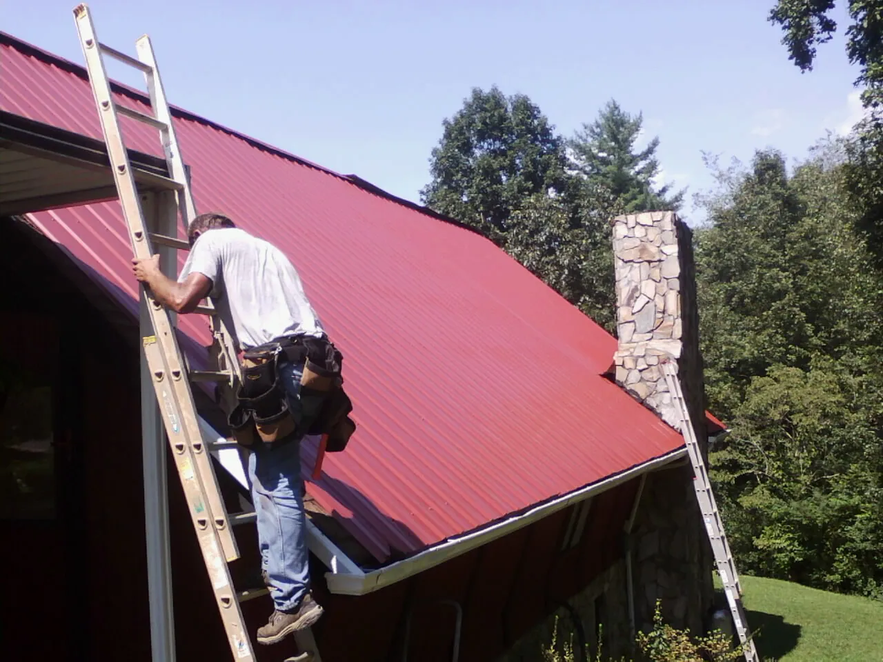 Roofer on a ladder beside a red metal roof and stone chimney — Soffit & Fascia Repair by Roofing Brothers in Matheson